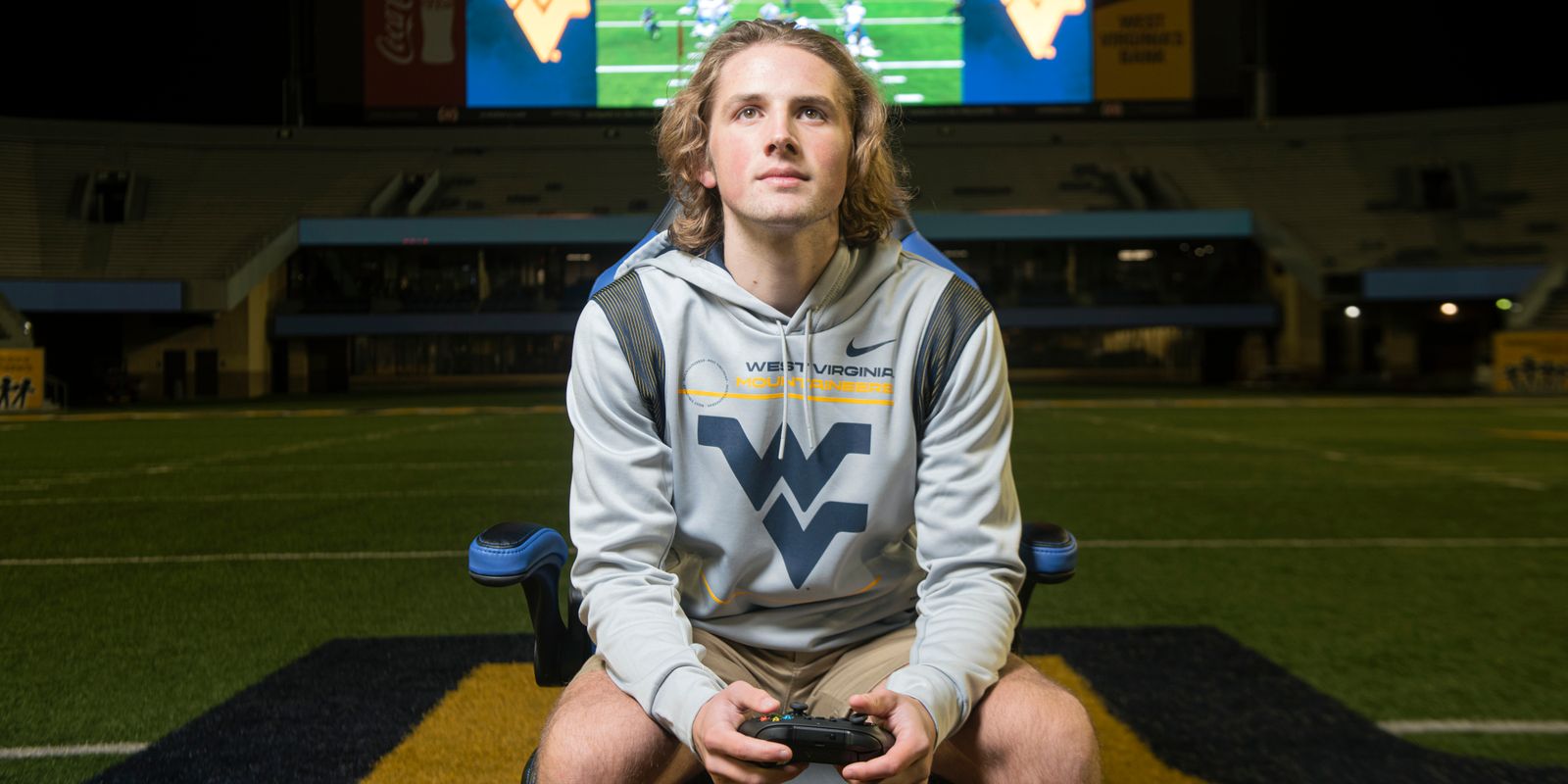 young man, seated, flying WV shirt, game controller, big screen in background