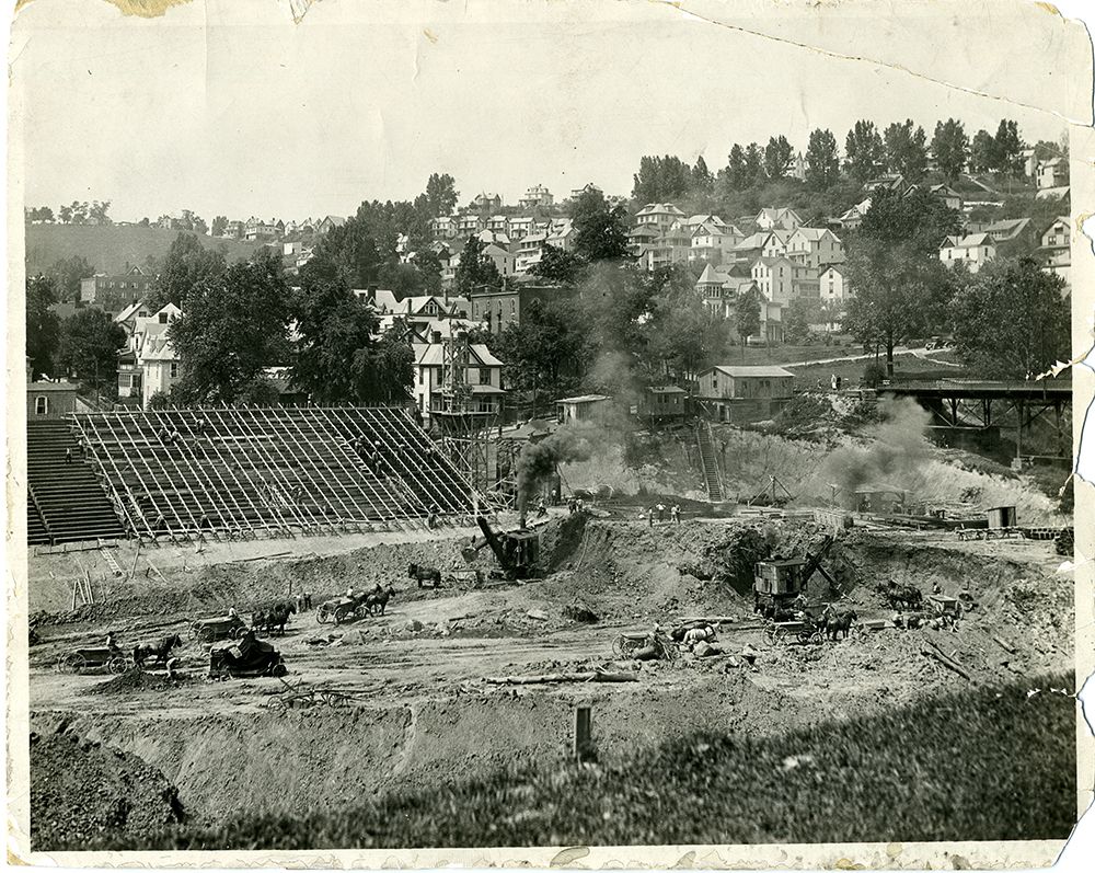 Old Mountaineer Field is under construction with excavators and wooden stands taking shape. A hill of homes and trees is in the background.