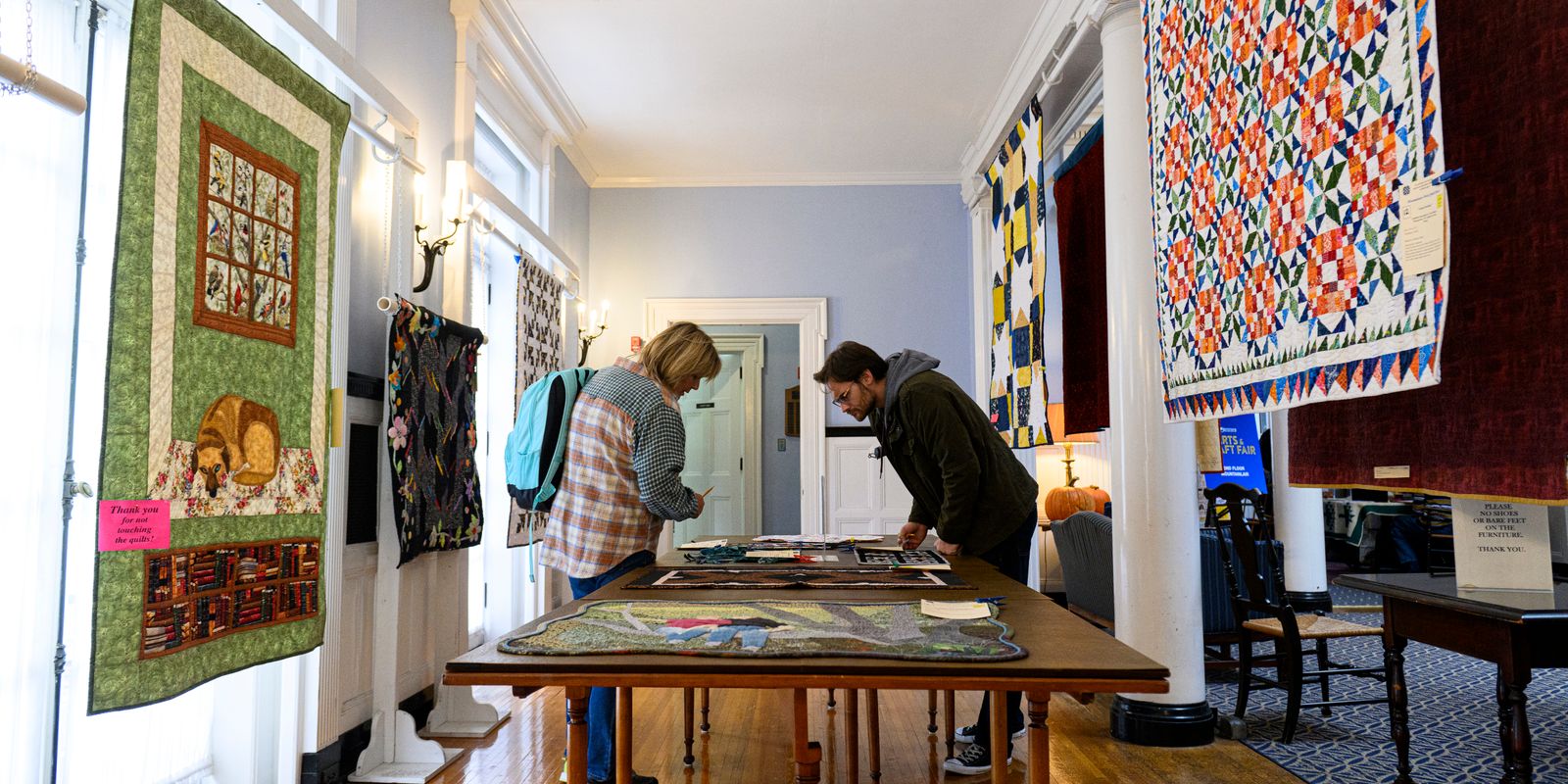 two people look at items on table, quilts hang in background