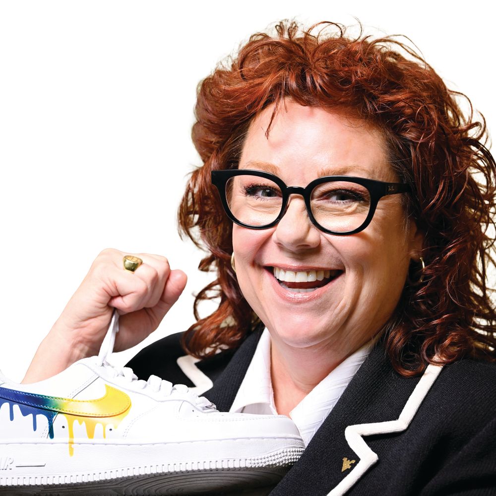 woman, red hair, glasses, holds sneaker with dripping gold and blue paint