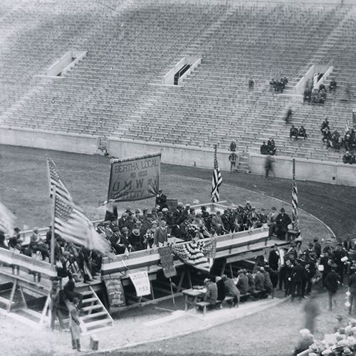 Photo of raised stage in old Mountaineer Field with a UMWA sign and people on the platform and empty stands on the side.