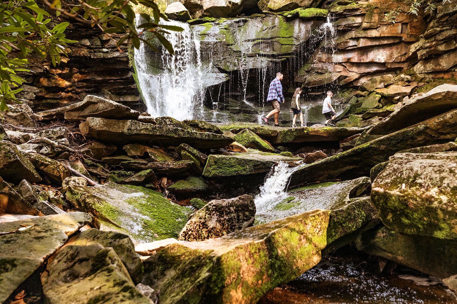 Photo of adult and two kids hiking a trail among rocks with a waterfall in the background at Blackwater Falls.