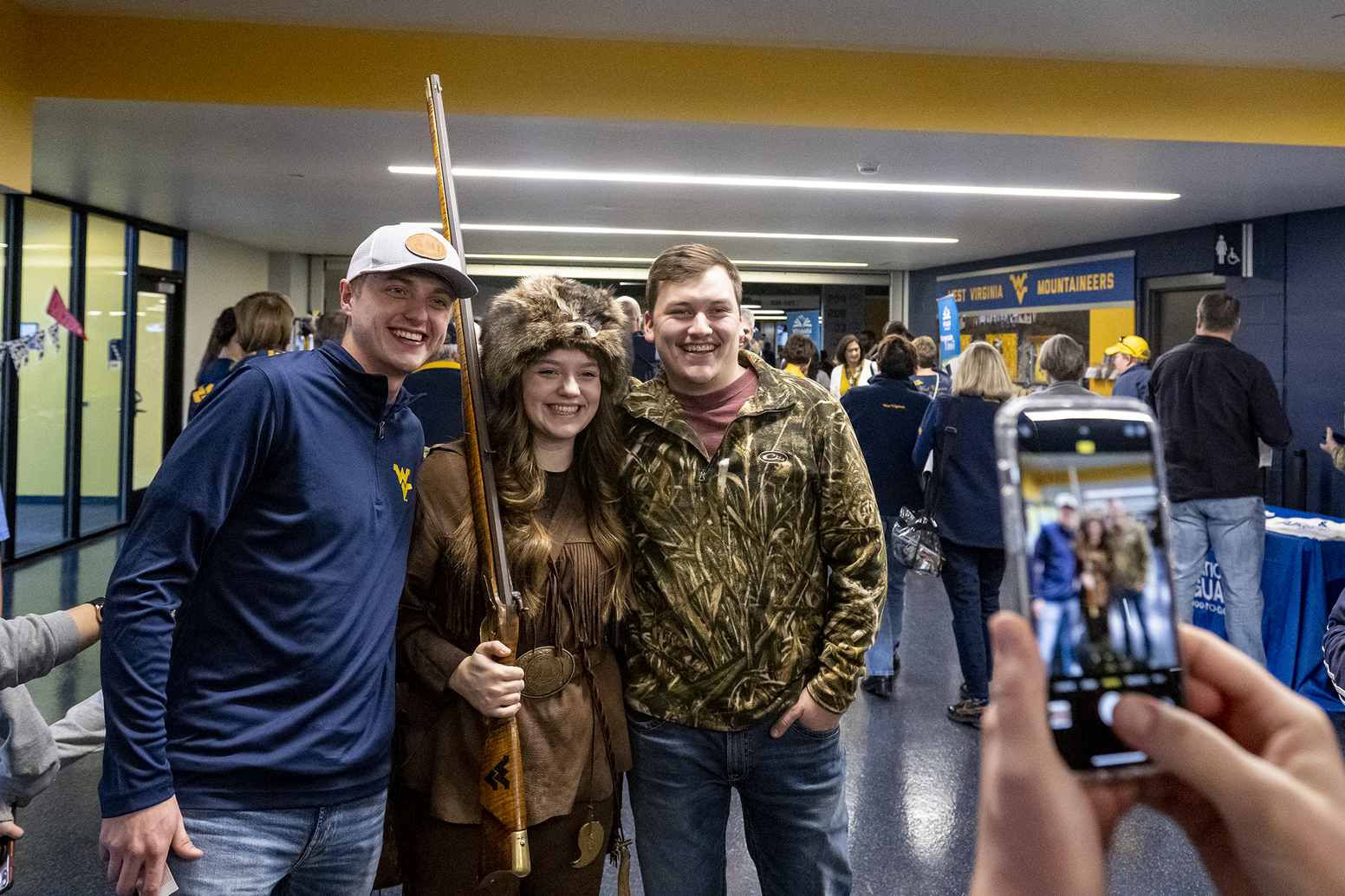 two young men flank woman in buckskins holding rifle, hands hold a cell phone taking a photo in the foreground