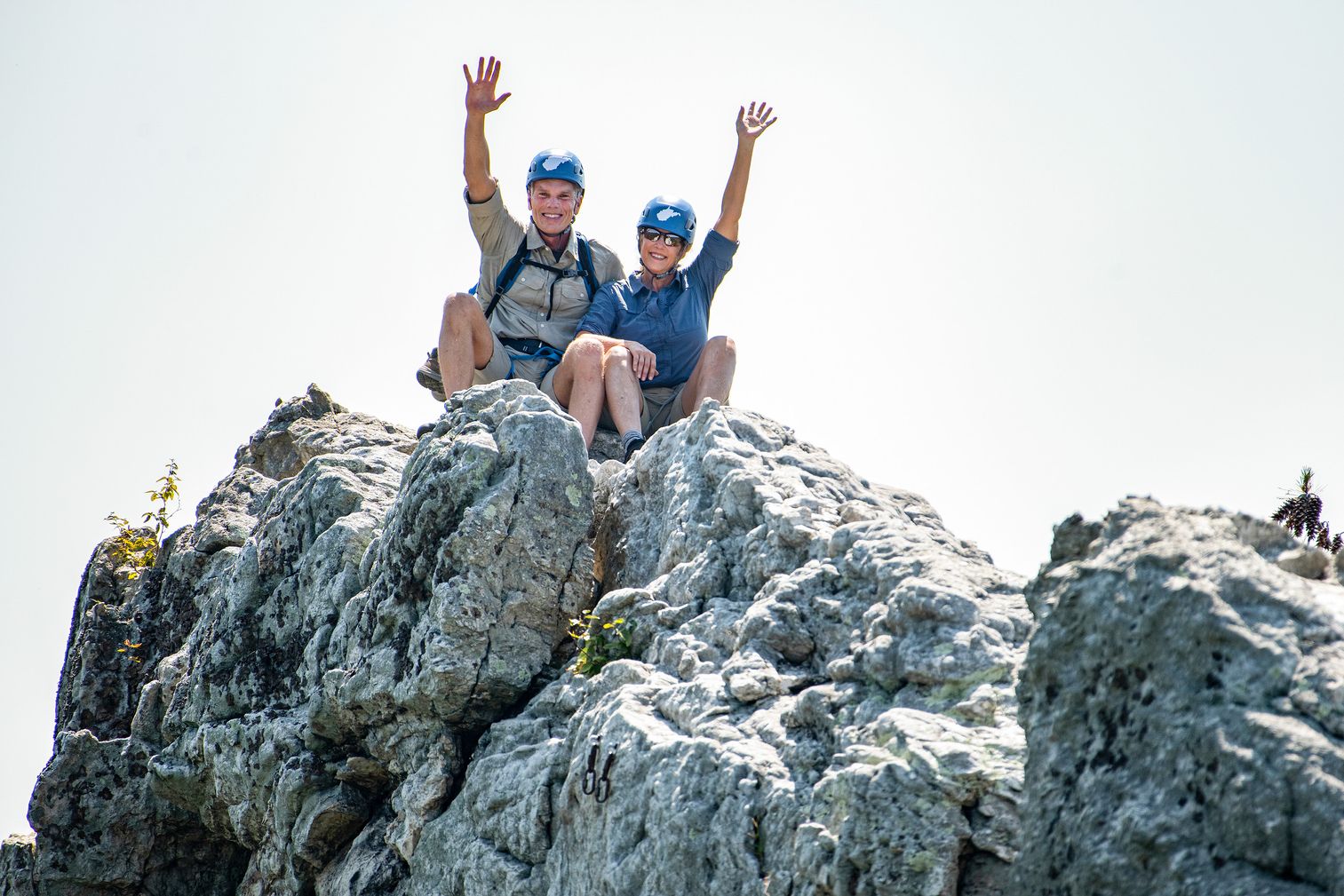 Brad and Alys Smith sit on a rocky summit with arms raised and wearing helmets.