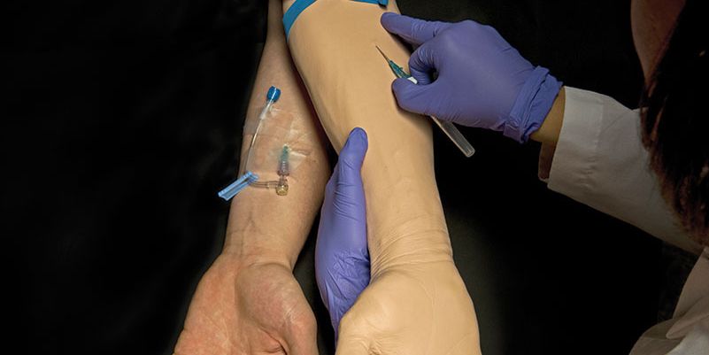 A student inserts a syringe into a manikin arm beside a human arm that has an IV inserted.