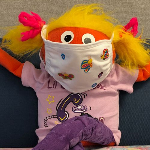 An orange puppet with yellow hair in pigtails is wearing a child's fabric mask.